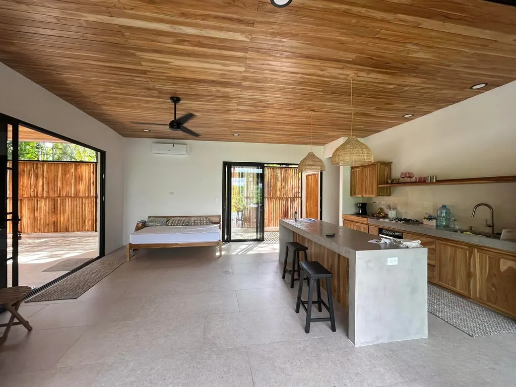 Open kitchen and dining in Casa Delicias with wood-slat ceiling, monolithic concrete island, and integrated bar seating.